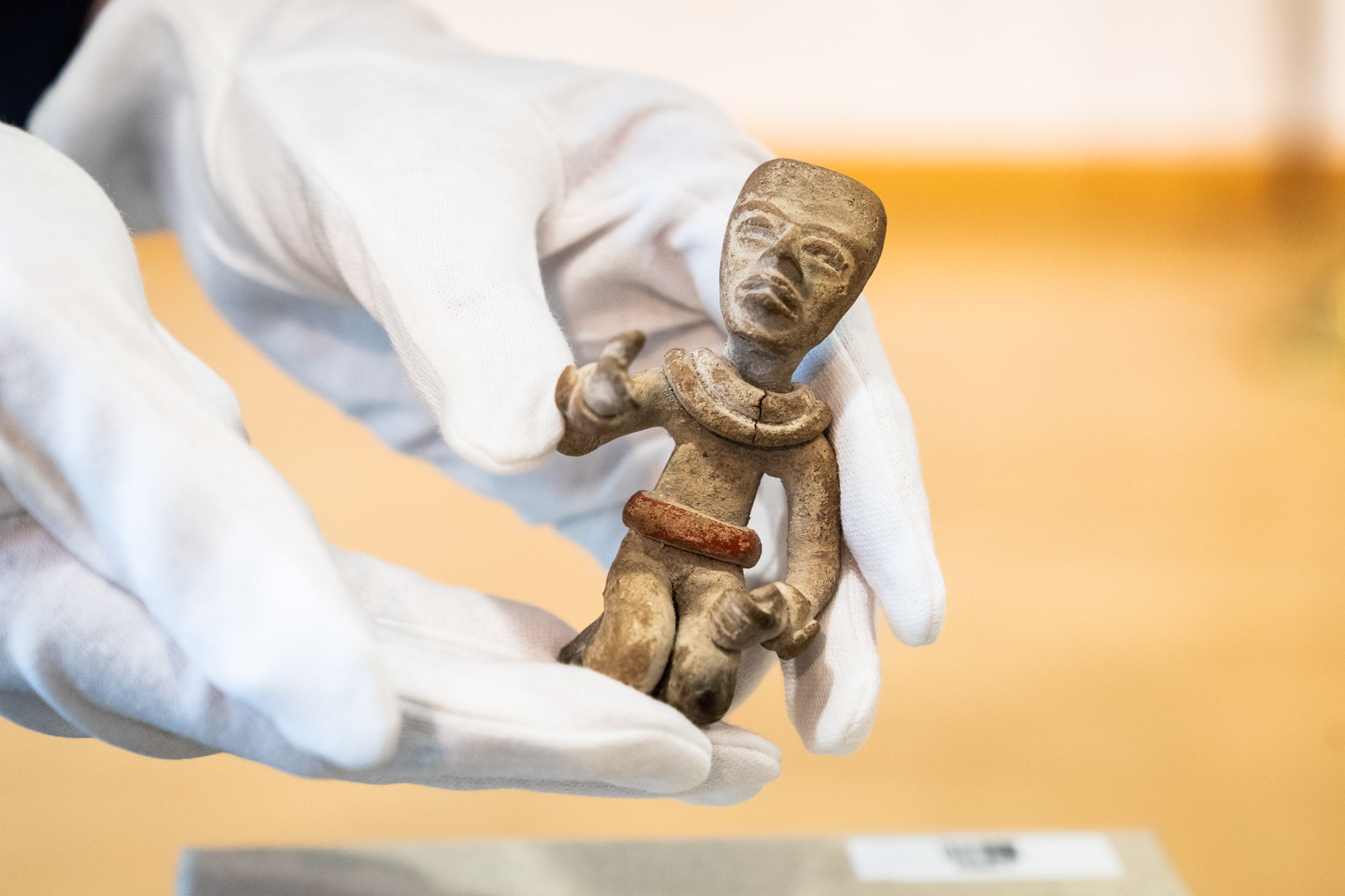 Zwei weiß behandschuhte Hände präsentieren eine kleine kniende Tonfigur eines Menschen, der einen Arm leicht erhebt, den anderen auf dem Oberschenkel ruhen lässt. (verweist auf: Maya-Artefakte nach Mexiko und Guatemala zurückgekehrt)